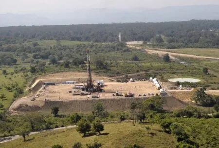 Aerial view of an oil drilling derrick on a fenced dirt pad with trucks and equipment scattered around in a rural landscape.