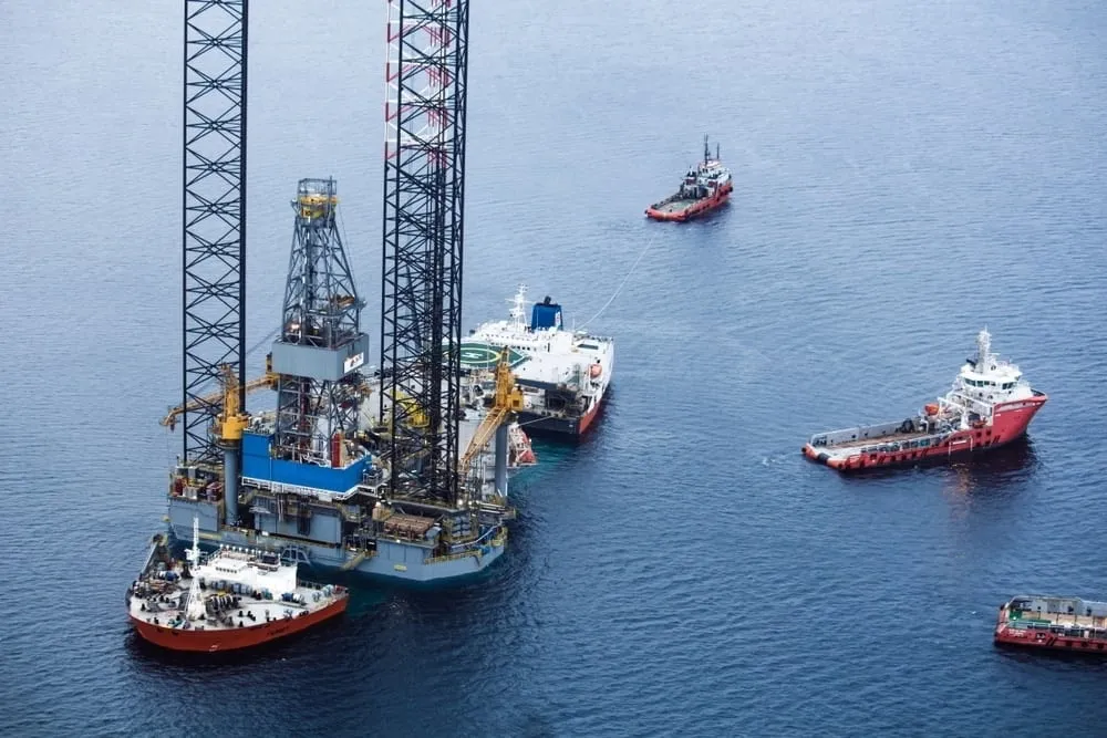 Offshore oil drilling rig with tall lattice legs, surrounded by tugboats and support vessels on calm blue water.