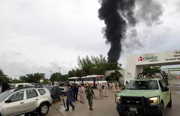 Smoke plume rising from a fire near a Pemex logistics terminal as people and vehicles gather on a roadway.