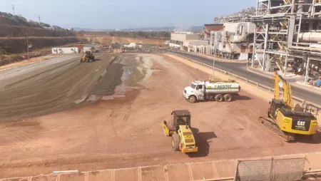Construction site with a dirt road being prepared for paving; a yellow road roller, an excavator, and a white tanker truck near a large industrial building on the right.