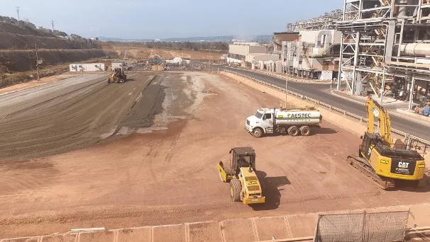 Construction site with a dirt road being prepared for paving; a yellow road roller, an excavator, and a white tanker truck near a large industrial building on the right.