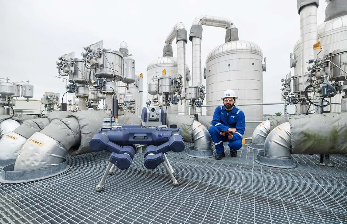Maintenance engineer in blue coveralls and hard hat kneels with a tablet beside a four‑legged industrial robot on a grated platform, surrounded by large pipes and tanks in a processing plant.