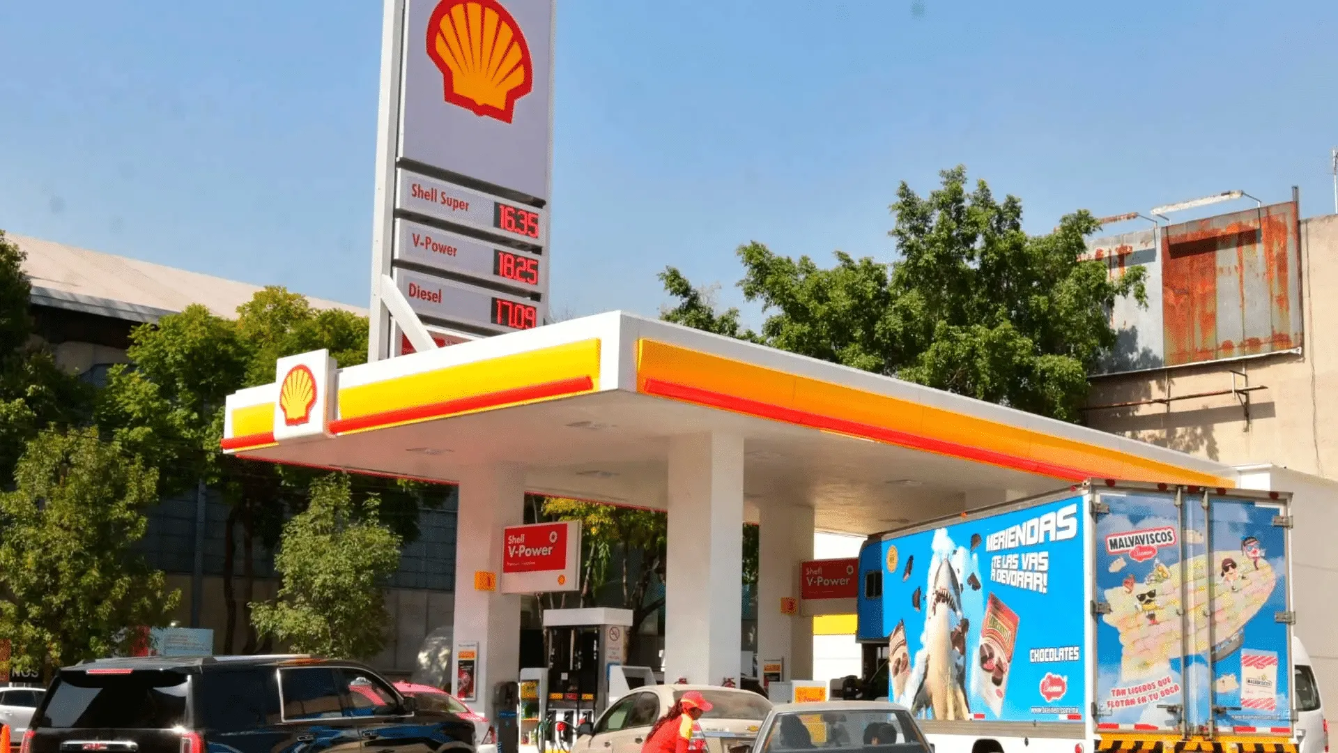 Shell gas station with a tall sign showing fuel prices and a white canopy with orange-yellow trim under a blue sky.
