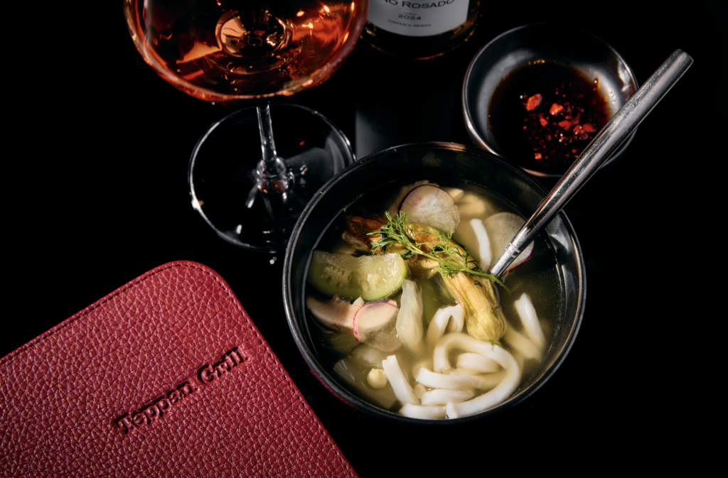 tepan grill 3 - Petróleo y Energía Bowl of noodle soup with greens and radish slices on a dark surface, with a glass of rosé nearby and a red textured item in the corner.