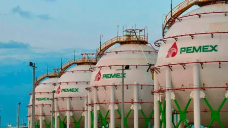 Row of large white storage tanks with red ladders and the PEMEX logo at an oil facility against a blue sky.