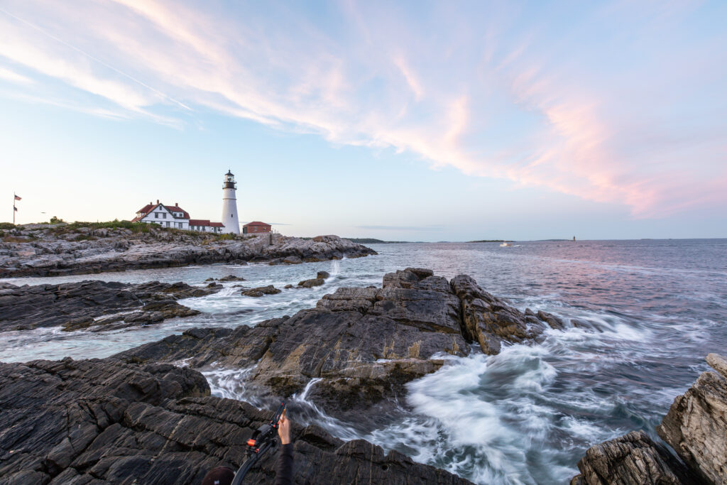 1 Maine - Faro en playa 7 - Petróleo y Energía Rocky coastline with a white lighthouse and red-roofed buildings under a pink sunset sky beside calm water.