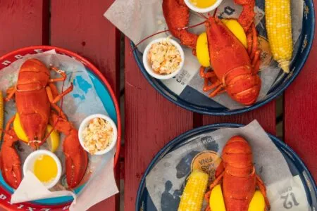 Two red lobsters served in blue baskets with corn on the cob and lemon wedges on a red table; small bowls of sauce and sides nearby.