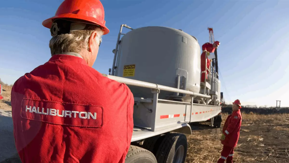 Back view of a Halliburton worker in red coveralls and a hard hat standing beside a large gray tank on a flatbed trailer at a rural worksite.