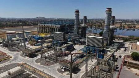 Large industrial power plant with tall smokestacks, cooling towers, and a dense network of pipes and buildings, set near a waterway.