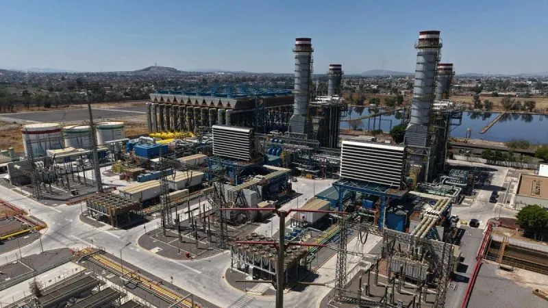 Large industrial power plant with tall smokestacks, cooling towers, and a dense network of pipes and buildings, set near a waterway.