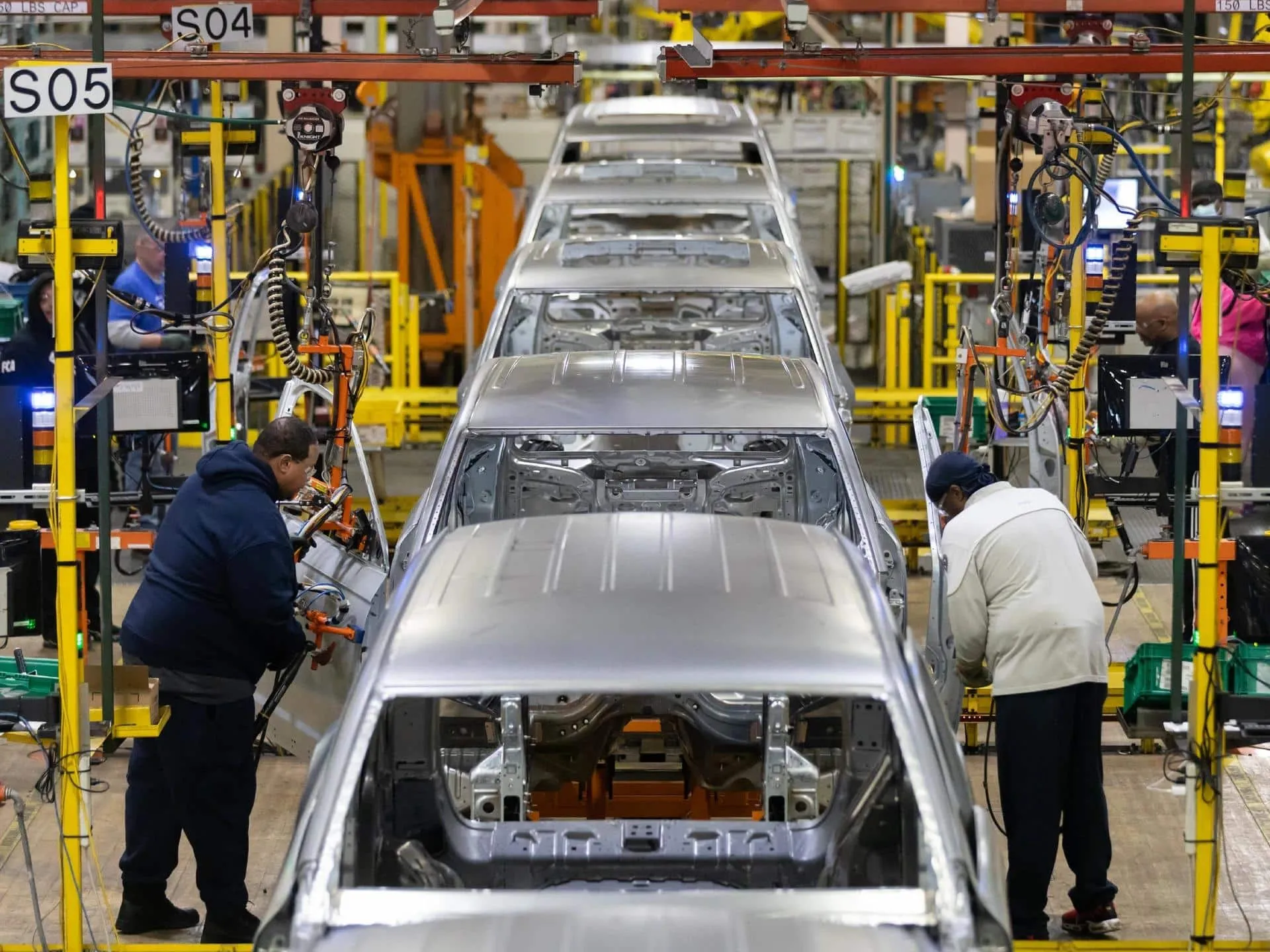 Factory assembly line with silver car bodies on an overhead conveyor, workers assembling panels in a busy automotive plant.