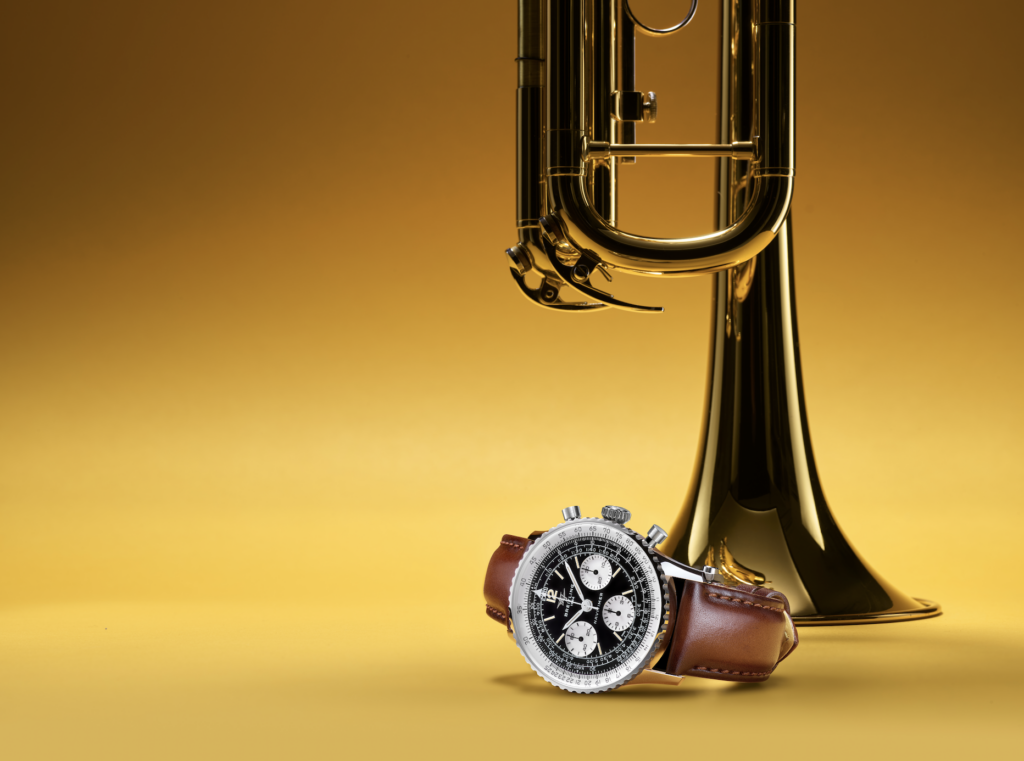 BREITLING JAZZ - Petróleo y Energía Close-up of a black-dial chronograph watch with a brown leather strap, placed beside a tall gold champagne flute on a gold gradient backdrop