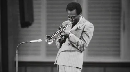 Jazz trumpeter in a pinstripe suit performing on stage, focused on blowing into the trumpet with a microphone nearby