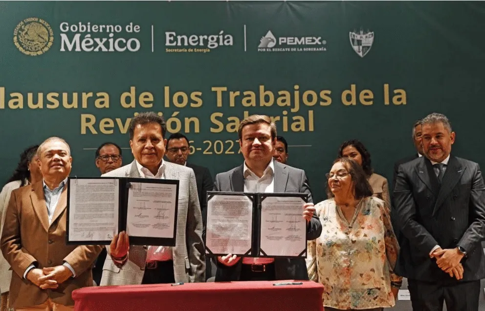 Group of officials signing documents during a salary review ceremony; two men hold open folders with signatures, others look on, behind them a green banner with logos and text in Spanish.