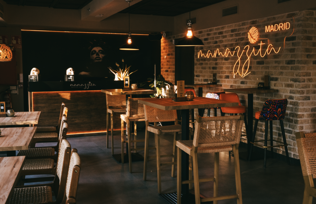 mamazzita madrid - Petróleo y Energía Dimly lit restaurant interior with wooden tables, woven chairs, brick walls, and a neon Madrid sign on the brick wall.