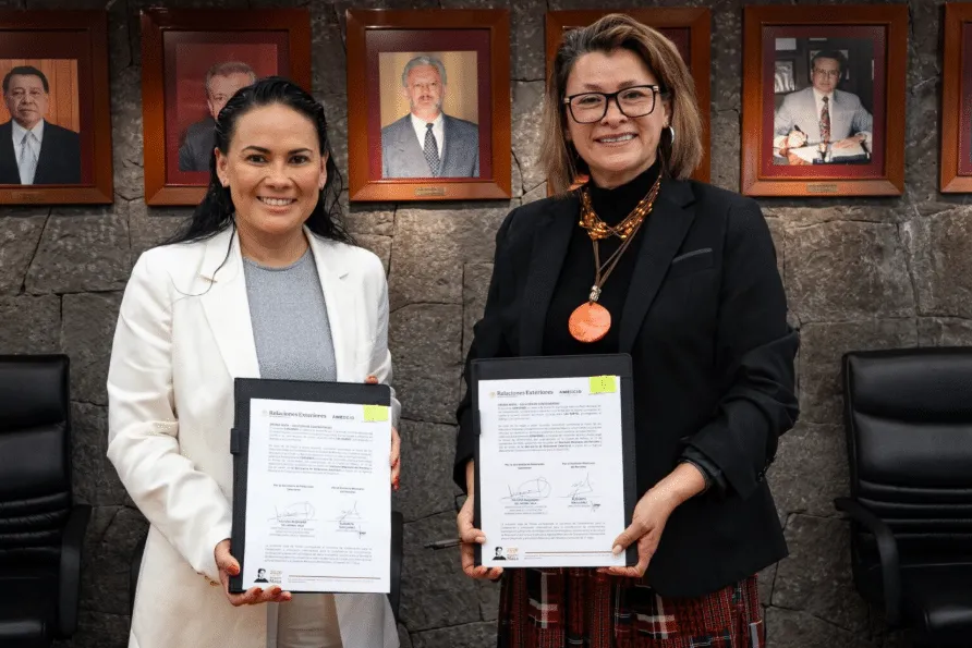 Two women in business attire pose with signed documents, standing against a stone wall with framed portraits behind them.