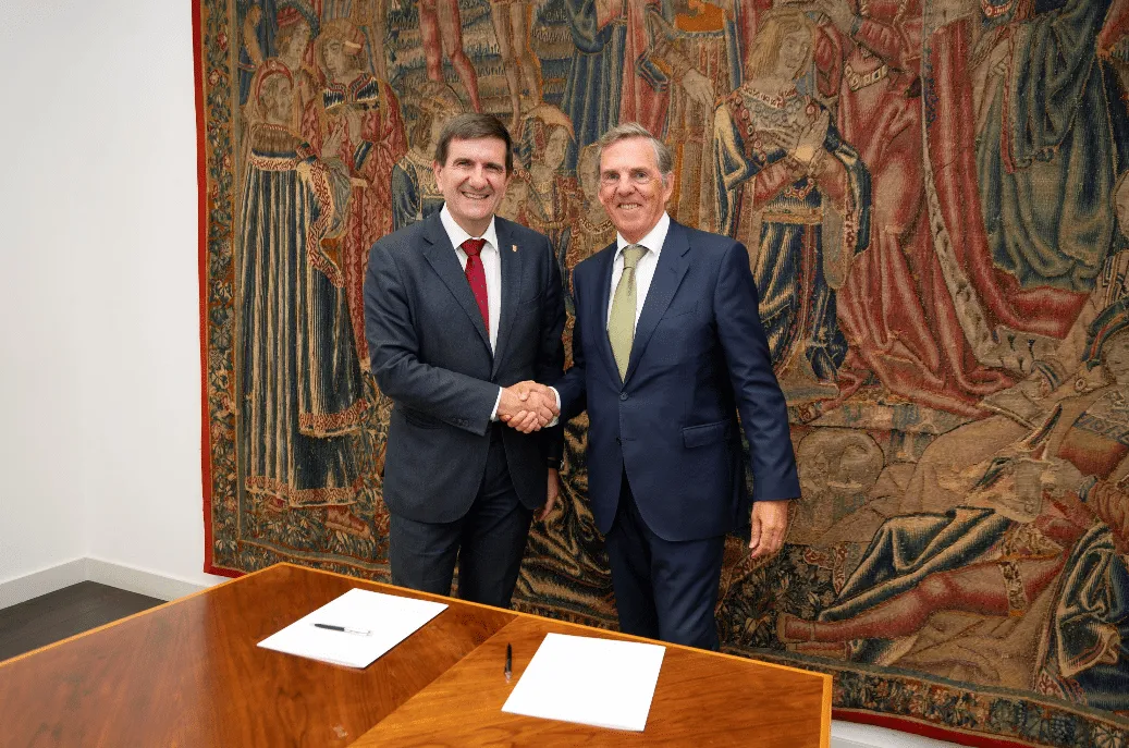 Two men in suits shake hands in front of a large tapestry, standing behind a wooden table with blank papers and pens.