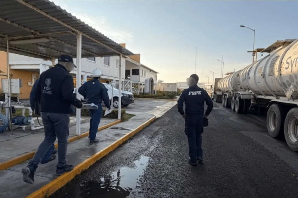 Emergency responders inspect a large fuel tanker parked at an industrial facility, walking along a wet, damaged road edge.