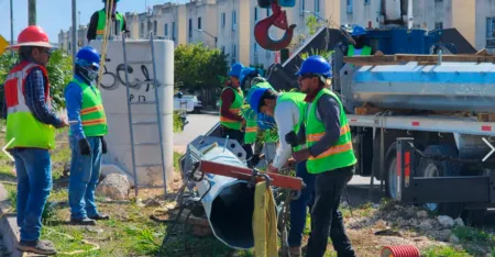 Construction workers wearing hard hats and high-visibility vests guide a large pipe at an urban construction site next to a truck.