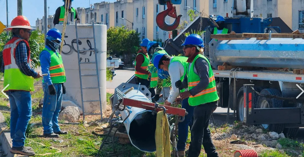 Construction workers wearing hard hats and high-visibility vests guide a large pipe at an urban construction site next to a truck.