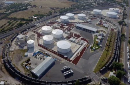 Aerial view of an industrial storage facility with multiple white cylindrical tanks connected by red pipelines within a circular rail loop around the site.