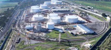 Aerial view of a large industrial facility with numerous white cylindrical storage tanks, rail tracks, and surrounding green fields.