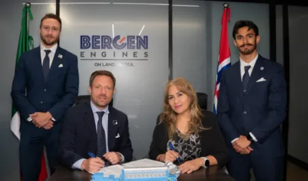 Four professionals in suits at a signing ceremony, two seated signing documents while a woman signs, with Bergen Engines logo and flags in the background.
