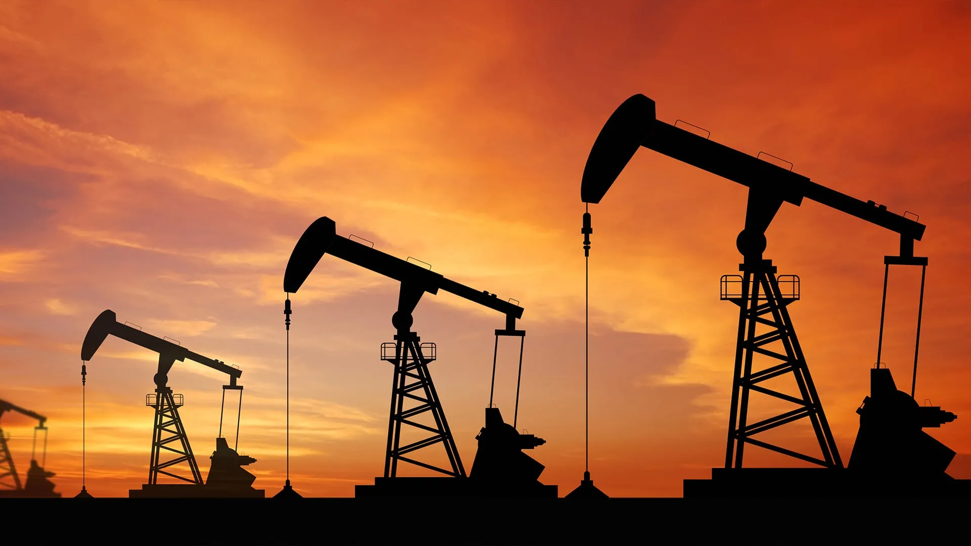Silhouettes of multiple oil pumpjacks at sunset with an orange sky.
