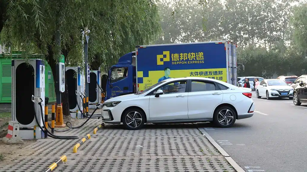 White sedan plugged into public EV charging stalls along a tree-lined street, with a blue Deppon Express delivery truck in the background.