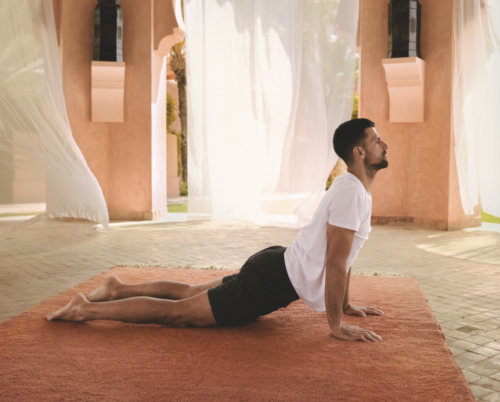 DJOKOVIC_AMANJENA 2 - Petróleo y Energía Man in a white t-shirt and black shorts performing a cobra yoga pose on an orange mat outdoors beneath a courtyard with flowing white drapes.