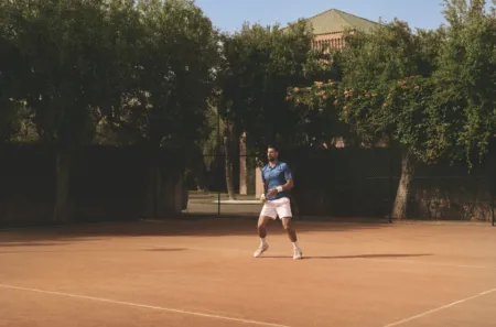 Male tennis player in a blue shirt and white shorts walks on a clay court, racket in hand, with a chain-link fence and trees in the background.