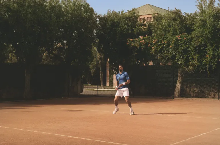 Male tennis player in a blue shirt and white shorts walks on a clay court, racket in hand, with a chain-link fence and trees in the background.