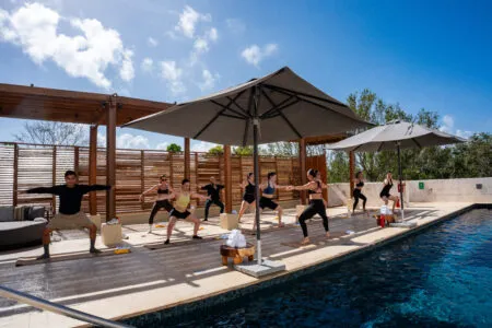 Group of people practicing yoga by a swimming pool under large umbrellas on a sunny outdoor deck.