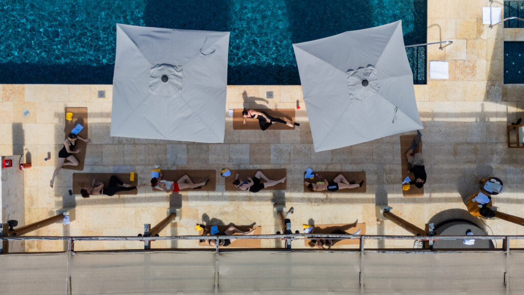default - Petróleo y Energía Aerial view of people sunbathing on lounge mats beside a blue pool under large gray umbrellas.