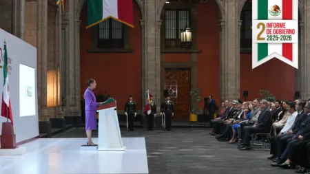 Woman in a purple suit delivers a speech at a podium in a formal Mexican government ceremony, with uniformed guards and seated dignitaries in the background.