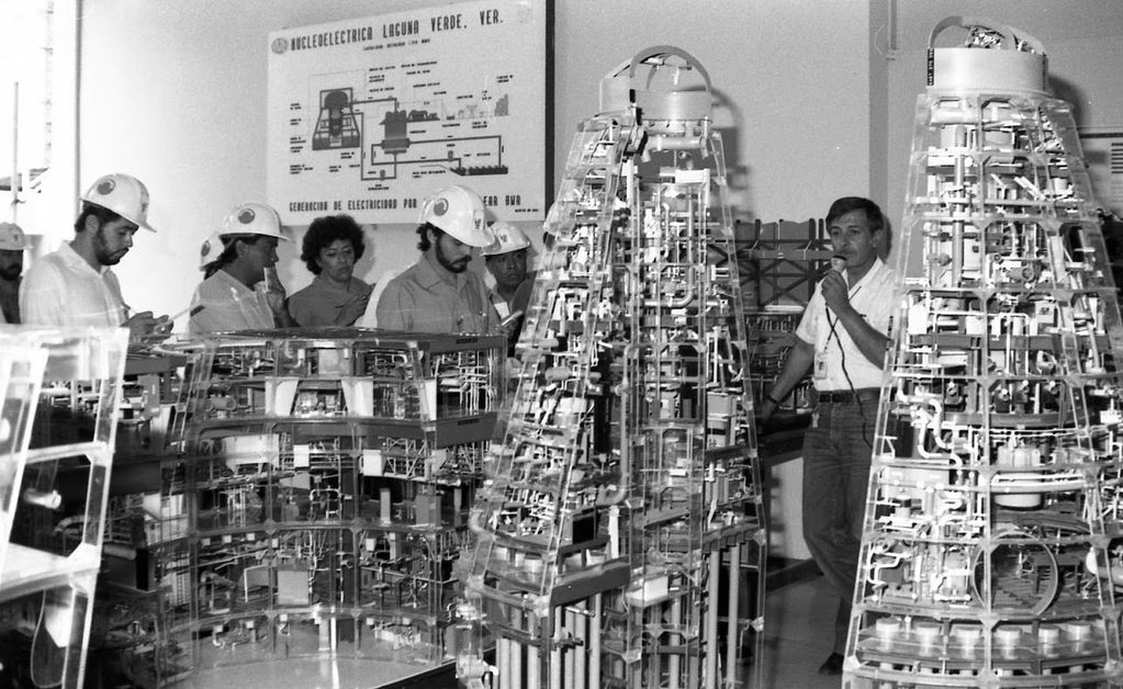 HFvyvjqXgAApP3g - Petróleo y Energía Group of engineers and technicians in hard hats examine tall modular electronic racks in a lab.