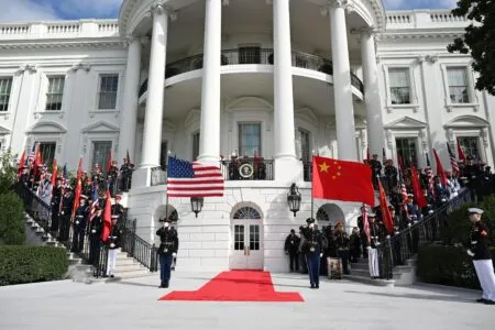 Military honor guards stand on a red carpeted staircase in front of a white neoclassical building, with US and Chinese flags displayed and visitors observing behind a railing.