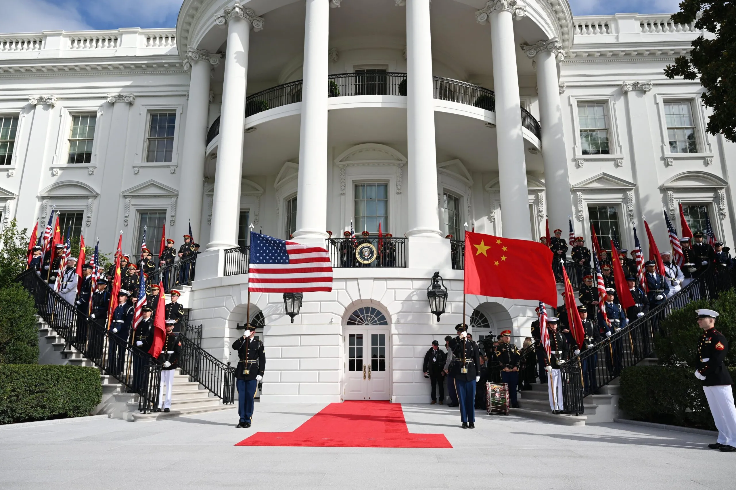 Military honor guards stand on a red carpeted staircase in front of a white neoclassical building, with US and Chinese flags displayed and visitors observing behind a railing.