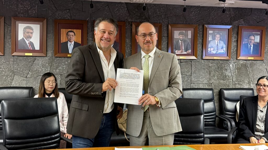 IMG_0543 - Petróleo y Energía Two men in suits shake hands while holding a document in a formal meeting room with framed portraits on a stone wall behind them, and seated people nearby.
