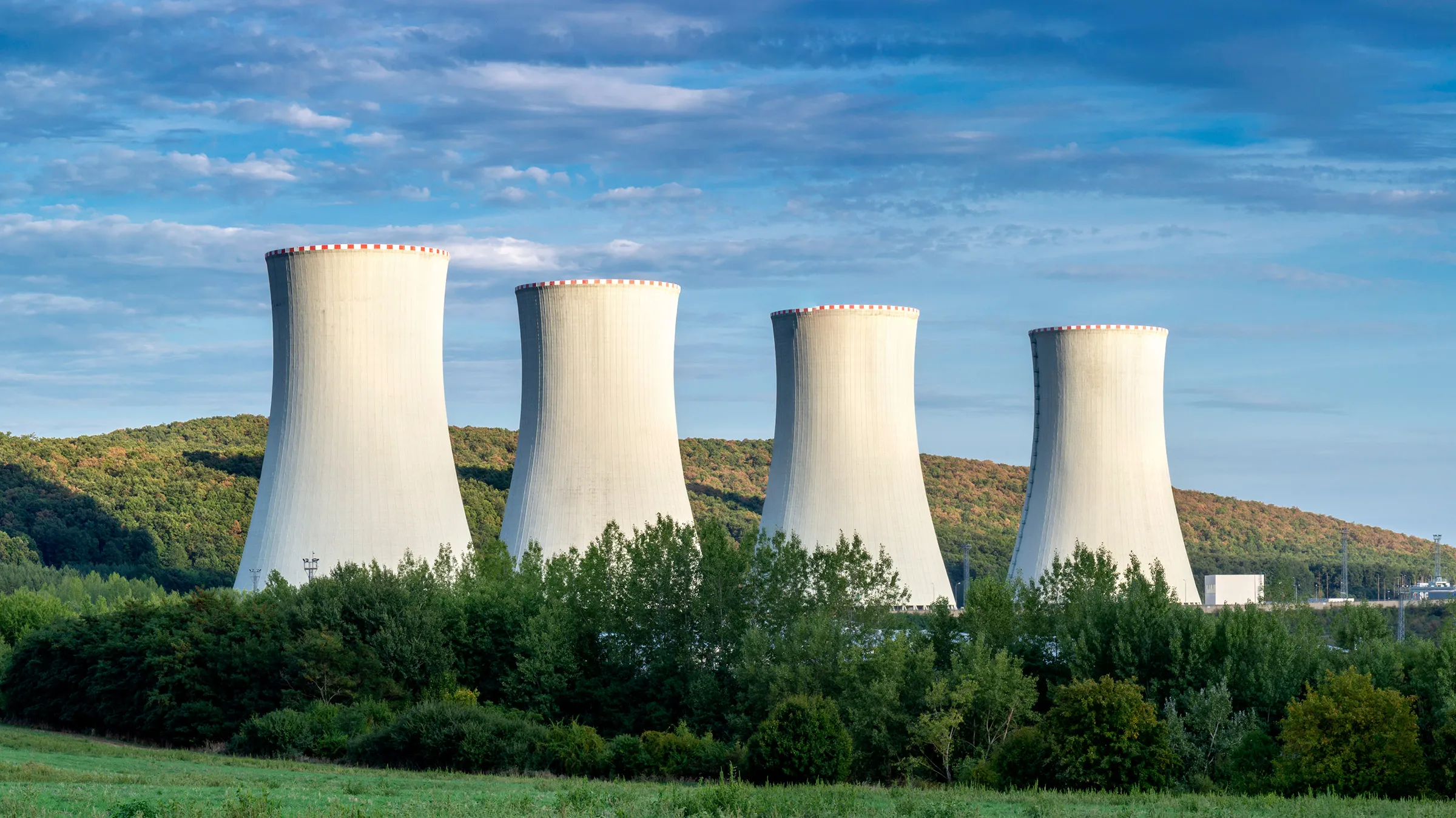 Four large cooling towers rise over a wooded landscape beneath a blue, partly cloudy sky.
