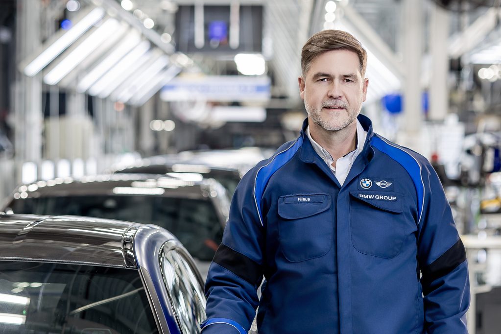 BMW Group technician named Klaus in blue coveralls standing beside a car on a bright assembly line floor.