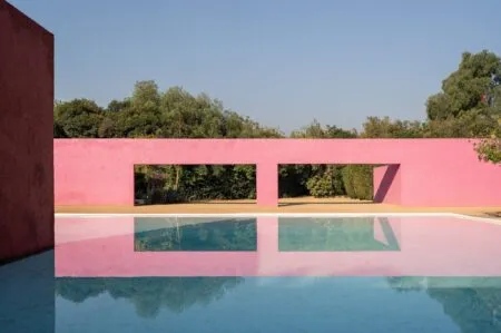 Pink stucco wall with rectangular openings overlooking a blue reflecting pool and green trees beyond, under a clear sky.