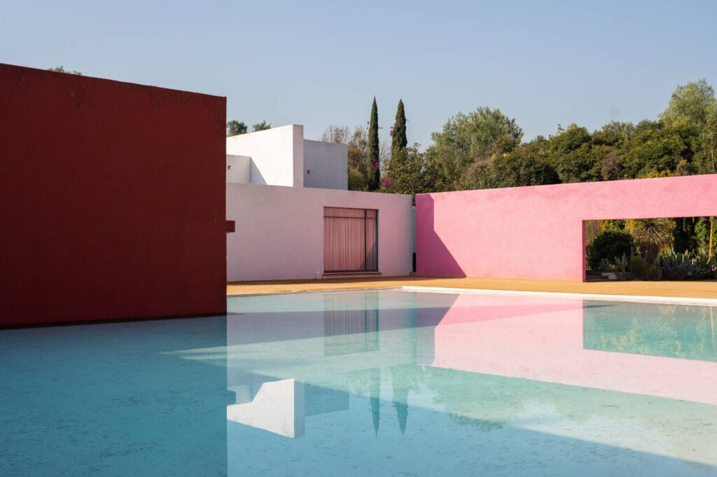LA CUADRA_FUNDACION ROMERO_PHOTO BY FABIAN MARTINEZ_2_LR - Petróleo y Energía Modern house exterior with pink and red stucco walls beside a reflecting pool under a clear blue sky.