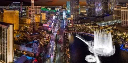 Aerial nighttime view of the Las Vegas Strip with neon hotels and the Bellagio fountain show on a lake.