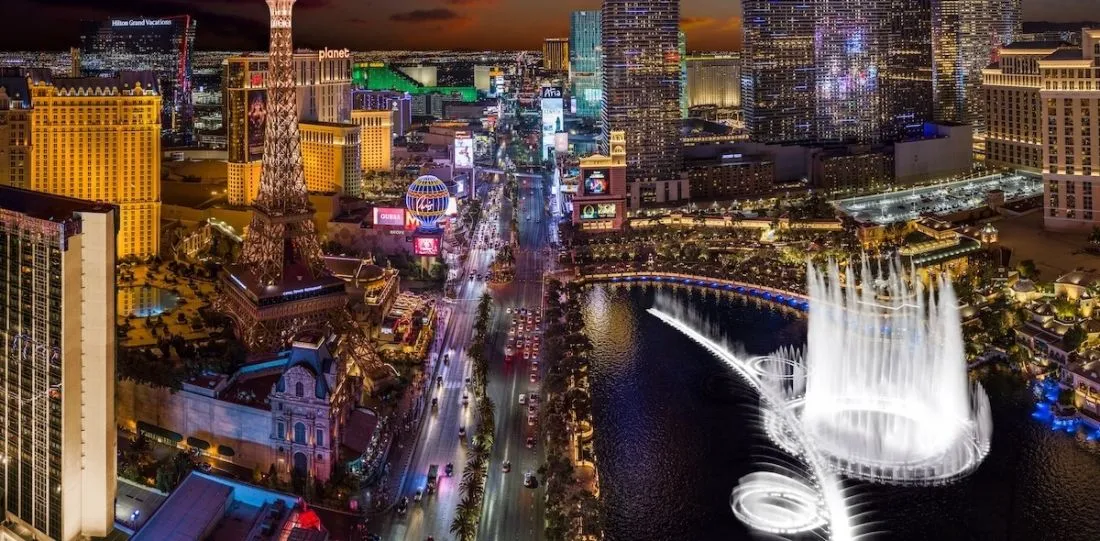 Aerial nighttime view of the Las Vegas Strip with neon hotels and the Bellagio fountain show on a lake.