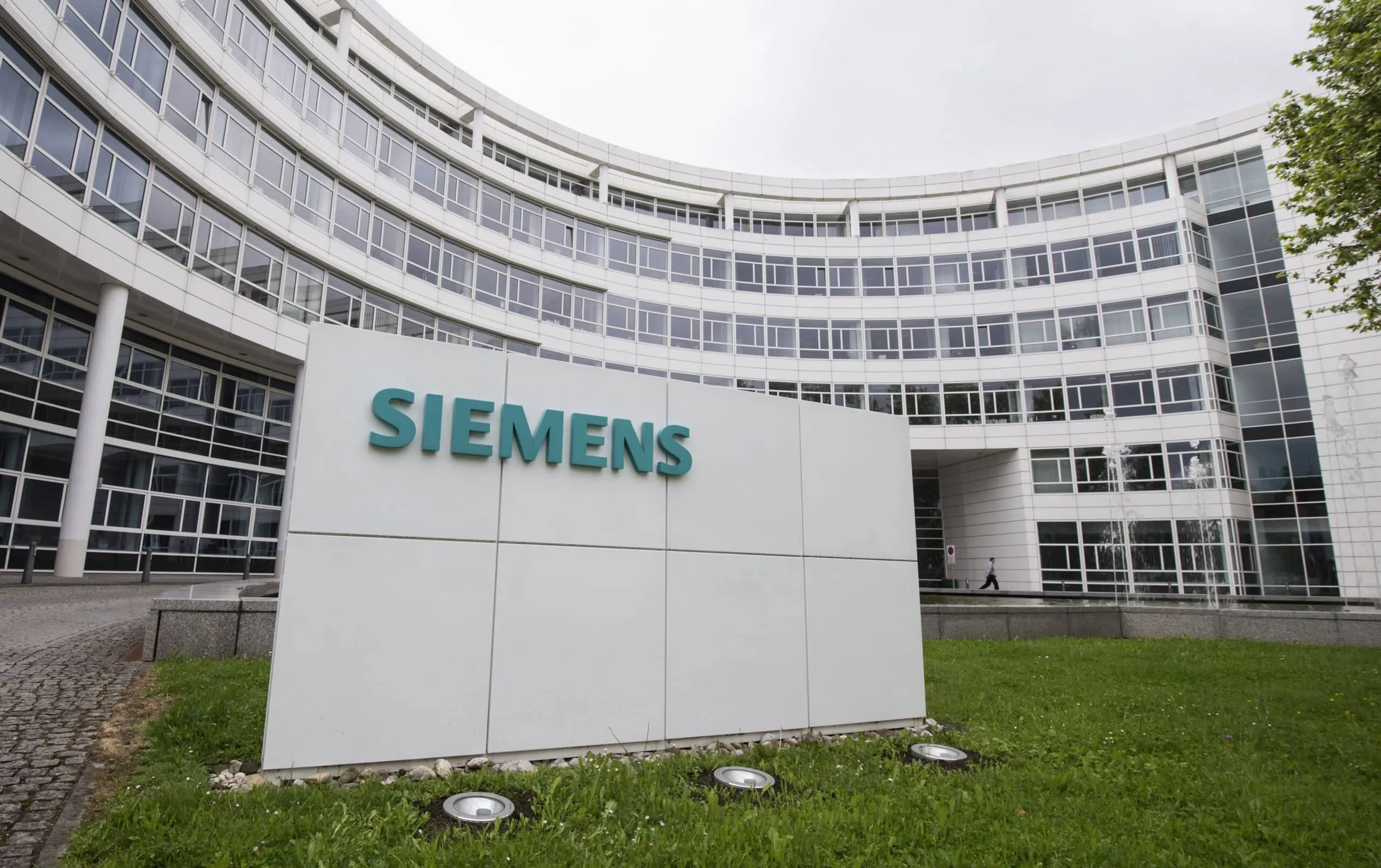 Curved modern Siemens office building with large glass windows; foreground shows a white sign reading SIEMENS in teal letters.