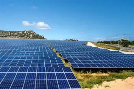 Large solar panel farm in a sunny landscape, rows of blue photovoltaic panels stretching toward distant hills under a clear blue sky.