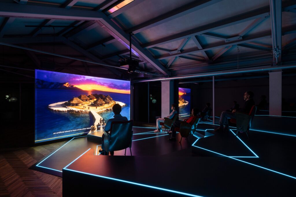 Osservatorio_Hito Steyerl - Petróleo y Energía Audience seated in a dark immersive exhibit watching a large curved screen showing a tropical island with huts and blue ocean.