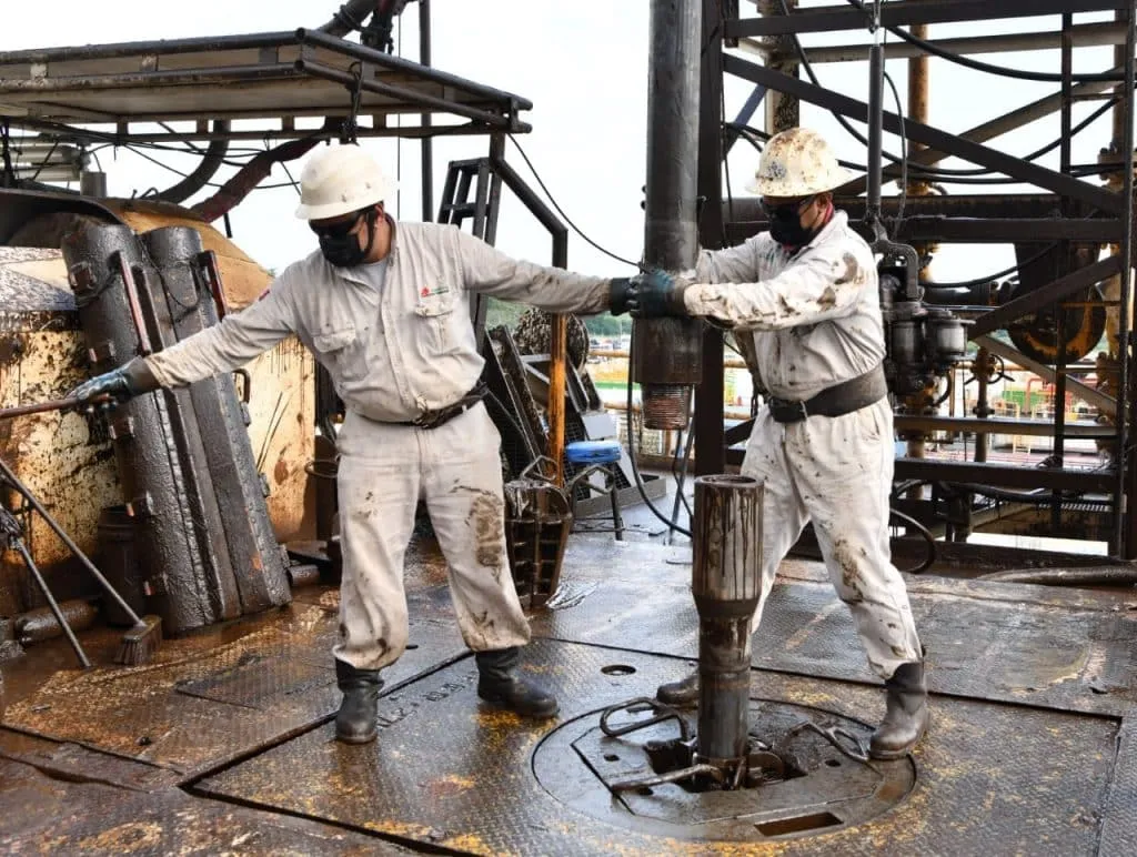 Two oilfield workers in hard hats and masks guide a drill pipe on a muddy, steel drilling platform.