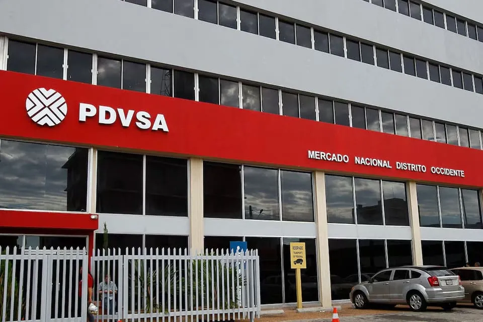 Exterior view of a PDVSA building with a bold red sign reading 'PDVSA' and 'Mercado Nacional Distrito Occidente,' glass windows, a metal fence, and parked cars nearby.